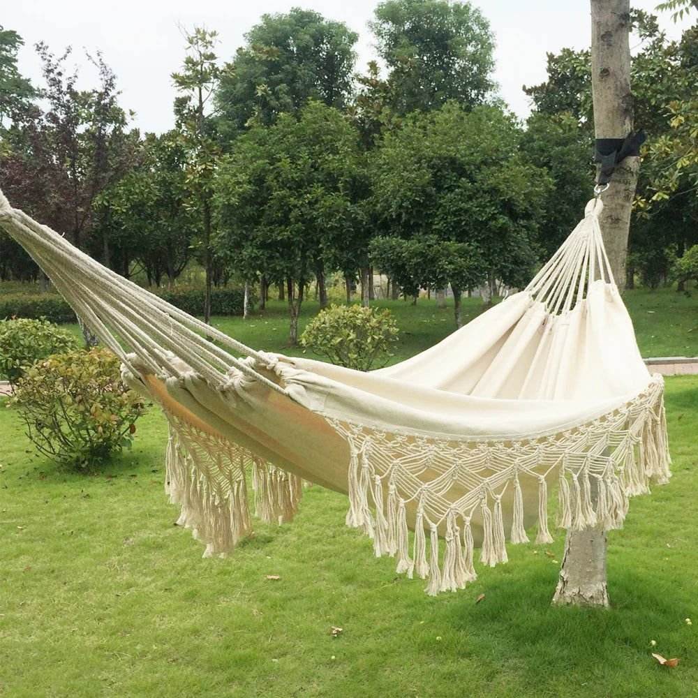 Double cotton hammock with macramé fringe hanging between trees in a green outdoor setting.
