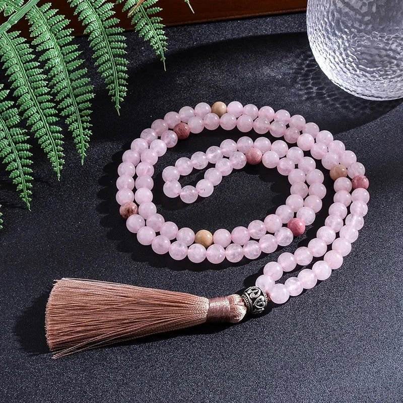 Rose Quartz Rhodonite Mala with pink tassel and OM charm on dark surface, surrounded by leaves.
